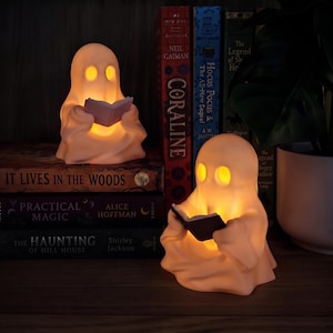 Könnte beinhalten: Zwei cremefarbene, geisterförmige LED-Lampen, die jeweils ein kleines offenes Buch halten. Die Geisterfiguren strahlen ein warmes, gelbes Licht aus ihren Augen und den Seiten der Bücher aus. Sie sind zwischen mehreren Büchern platziert.