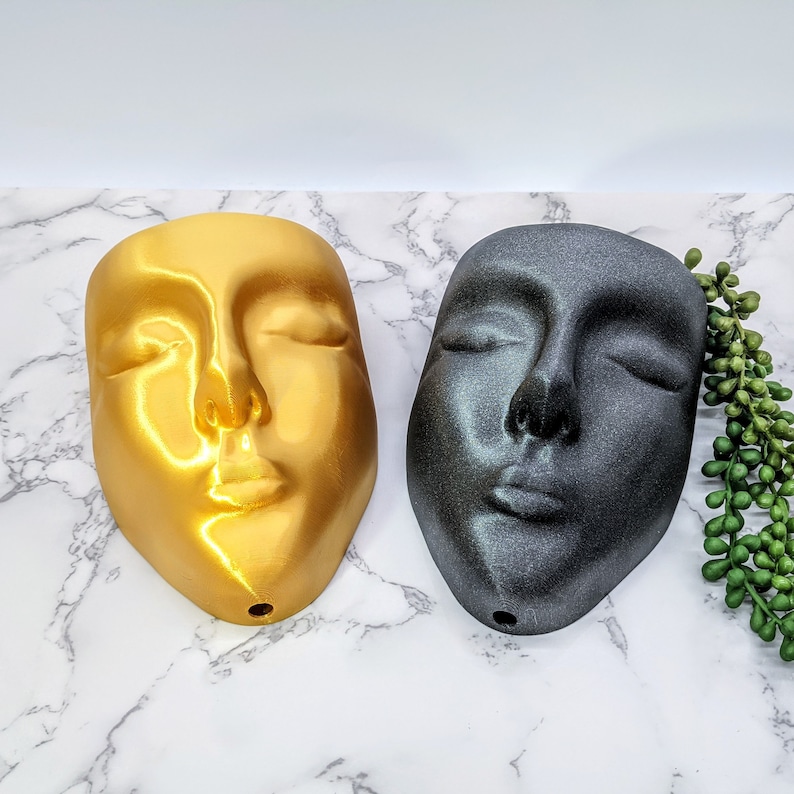 May include: Two face-shaped planters, one gold and one black, with closed eyes and a slightly parted mouth. The planters are on a white marble surface with a green plant in the background.