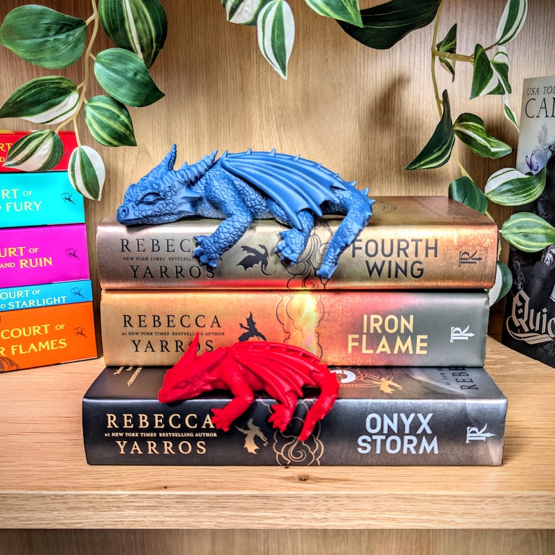 Puede incluir: Una figura de drag&oacute;n azul descansa sobre una pila de libros, incluyendo "Fourth Wing", "Iron Flame" y "Onyx Storm". Una figura de drag&oacute;n rojo est&aacute; en el libro inferior. Los libros est&aacute;n en una estanter&iacute;a de madera con plantas verdes.