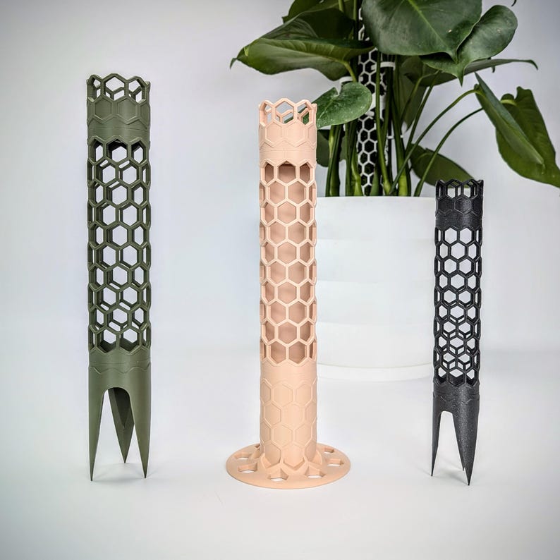 May include: Three plant supports made of plastic with a honeycomb design. The supports are in green, pink, and black. The supports are designed to help plants grow upright.