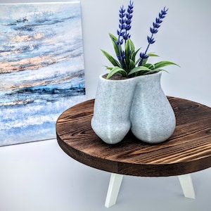 Puede incluir: Un macetero de cerámica blanca con una forma única y abstracta, que presenta dos secciones redondeadas. El macetero está lleno de flores de lavanda y se encuentra sobre una mesa de madera marrón oscuro con patas blancas.