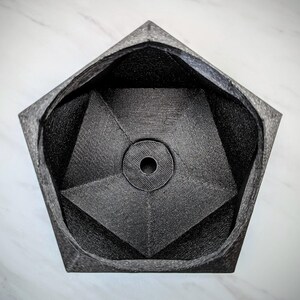 May include: A black, geometric, pentagonal planter with a hole in the center. The planter is made of a textured material and has a smooth, rounded interior.
