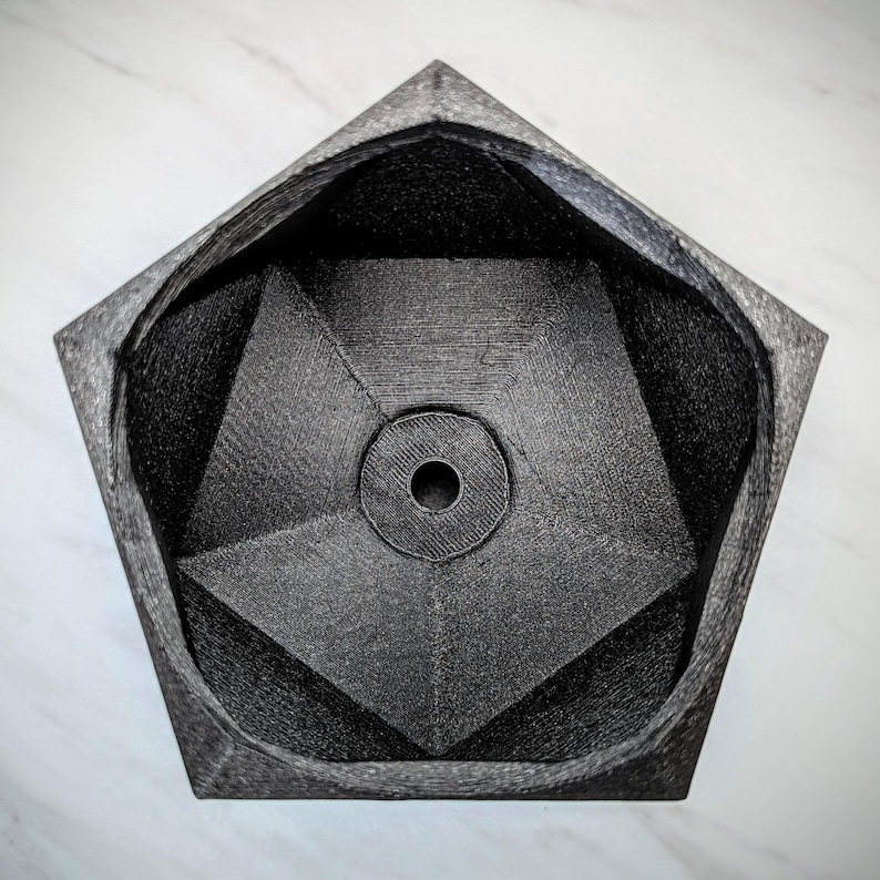 May include: A black, geometric, pentagonal planter with a hole in the center. The planter is made of a textured material and has a smooth, rounded interior.