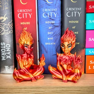 May include: Two red and orange figurines of a woman with flame-like hair and body details, seated in a meditative pose. The figurines are in front of a bookshelf with books titled "Crescent City" and "Onyx Storm".