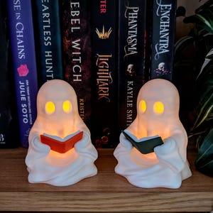 May include: Two white ghost-shaped lamps with glowing yellow eyes, each holding a small book. One ghost holds an orange book, and the other holds a dark green book. The figures are positioned on a wooden surface in front of a row of hardcover books.