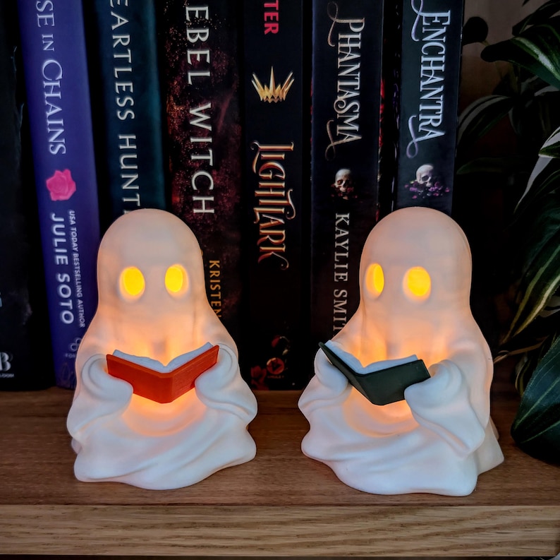 May include: Two white ghost-shaped lamps with glowing yellow eyes, each holding a small book. One ghost holds an orange book, and the other holds a dark green book. The figures are positioned on a wooden surface in front of a row of hardcover books.