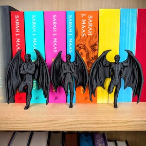 Puede incluir: Tres figuras negras impresas en 3D de personajes masculinos alados frente a una fila de libros coloridos de Sarah J. Maas. Cada figura presenta grandes alas detalladas, una complexi&oacute;n musculosa y poses distintas, todo en un acabado negro mate.