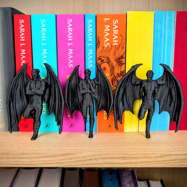 Puede incluir: Tres figuras negras impresas en 3D de personajes masculinos alados frente a una fila de libros coloridos de Sarah J. Maas. Cada figura presenta grandes alas detalladas, una complexi&oacute;n musculosa y poses distintas, todo en un acabado negro mate.