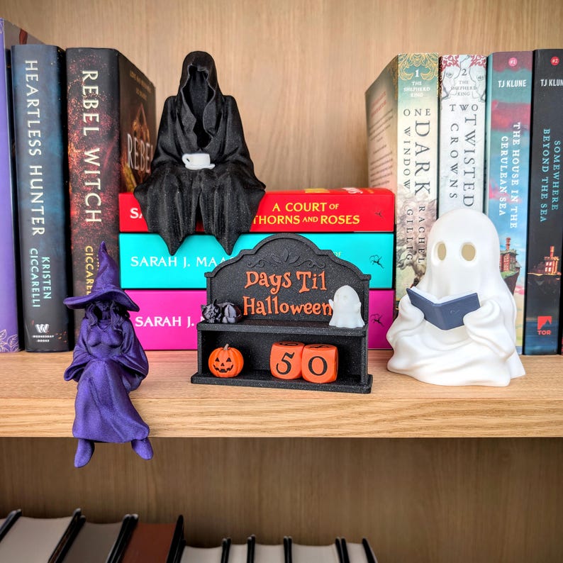 May include: A black hooded figure holding a white cup, a purple witch figurine, a white ghost reading a book, and a black shelf with the text Days Til Halloween, a small pumpkin, and two orange numbered blocks sitting on a wooden bookshelf.