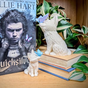 Könnte beinhalten: Zwei weiße Keramik-Fuchsfiguren, eine große und eine kleine, jeweils mit einem blauen Schmetterling auf der Nase. Die Figuren stehen auf Büchern, mit einem Buch und einer Pflanze im Hintergrund.