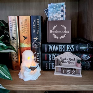 May include: A white ghost-shaped lamp holding a small book, positioned next to three Rebecca Yarros books, a brown box labeled Bookmarks, a stack of books including Powerless, and a small shelf display with the text Books Read This Year and 152.