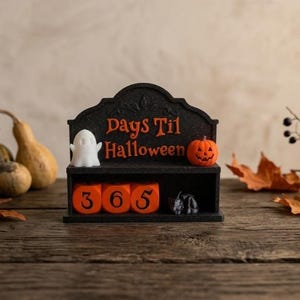 May include: A black countdown calendar with the orange text Days Til Halloween, featuring a white ghost figure on the left and an orange jack-o'-lantern on the right. The base contains three orange blocks displaying the number 365 in black.