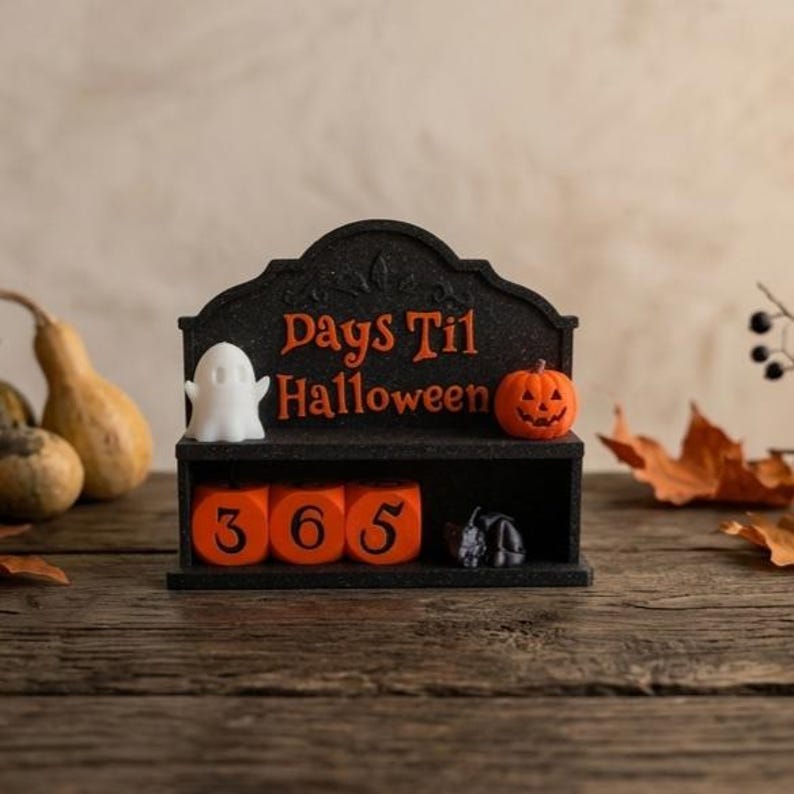 May include: A black countdown calendar with the orange text Days Til Halloween, featuring a white ghost figure on the left and an orange jack-o'-lantern on the right. The base contains three orange blocks displaying the number 365 in black.