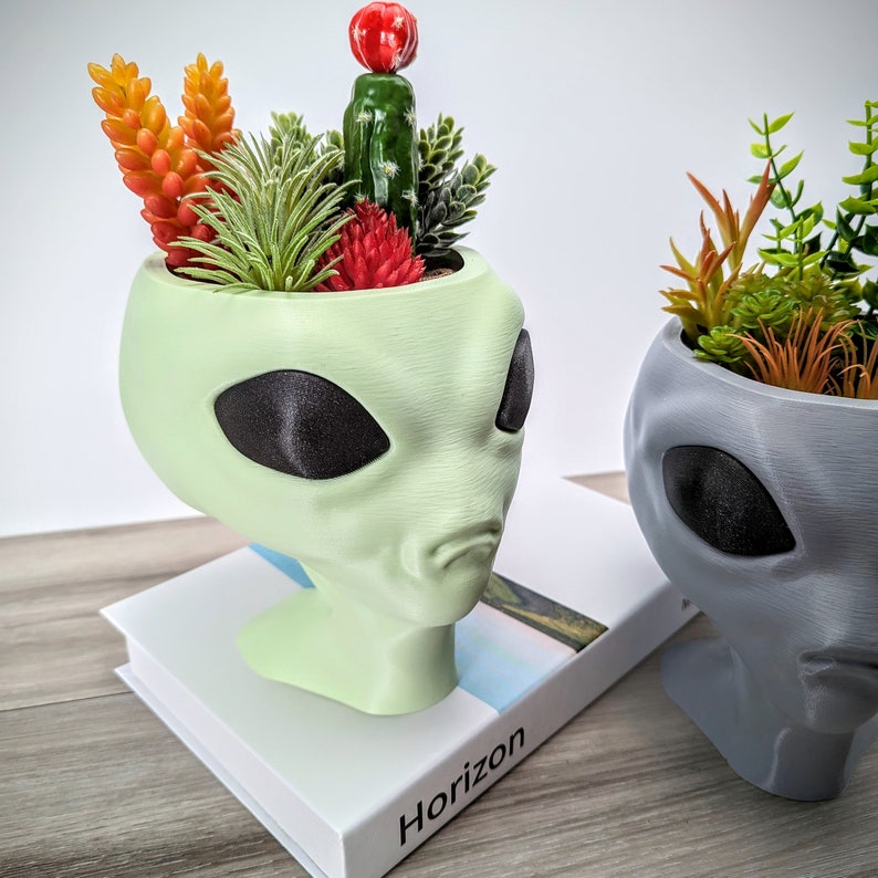 May include: Two green and gray alien head planters with succulents and cacti. The green alien planter is on a white book with the word "Horizon" printed on the cover. The gray alien planter is on a wooden surface.