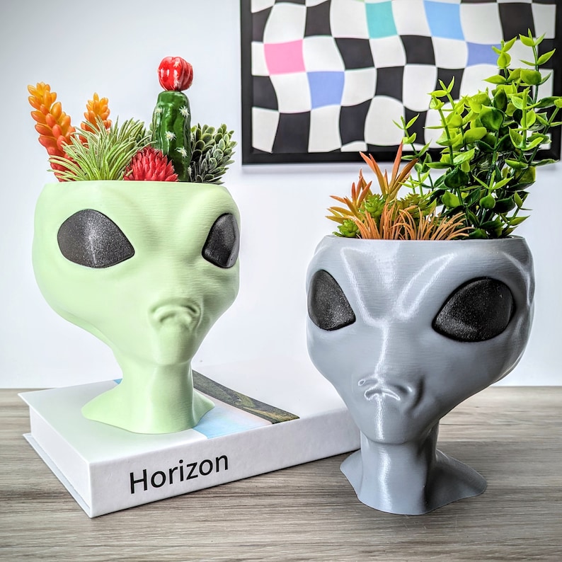 May include: Two alien head planters, one green and one gray, with different succulents planted inside. The green planter is on a book with the word "Horizon" printed on the cover. The gray planter is on a wooden surface.