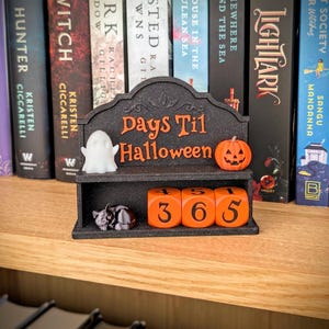 Può includere: Un calendario per il conto alla rovescia nero con la scritta arancione "Days Til Halloween", dotato di una figura di fantasma bianco, una zucca arancione, una piccola statuetta di gatto nero e tre cubi numerati arancioni con le cifre 4, 5, 1, 3, 6 e 5.