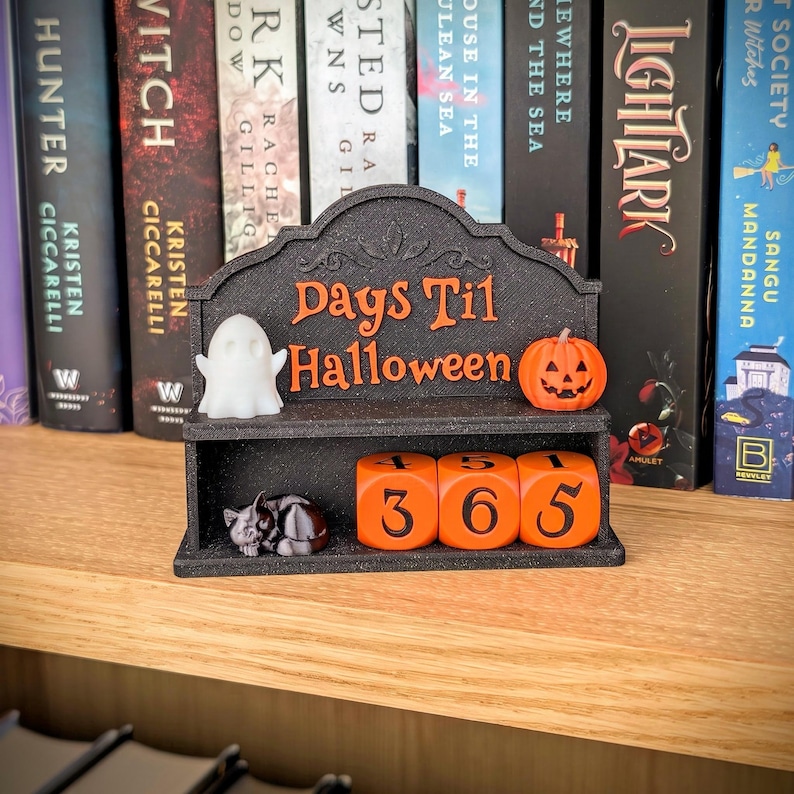 May include: A black countdown calendar with the orange text "Days Til Halloween" featuring a white ghost figure, an orange jack-o'-lantern, a small black cat figurine, and three orange numbered cubes displaying the digits 4, 5, 1, 3, 6, and 5.