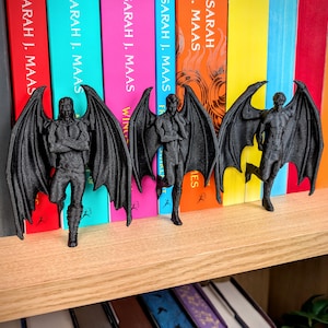 Puede incluir: Tres figuras negras impresas en 3D de personajes masculinos alados en diferentes poses. Cada figura presenta grandes y detalladas alas similares a las de un murci&eacute;lago y se encuentra de pie. Las figuras est&aacute;n sobre un estante de madera frente a libros.
