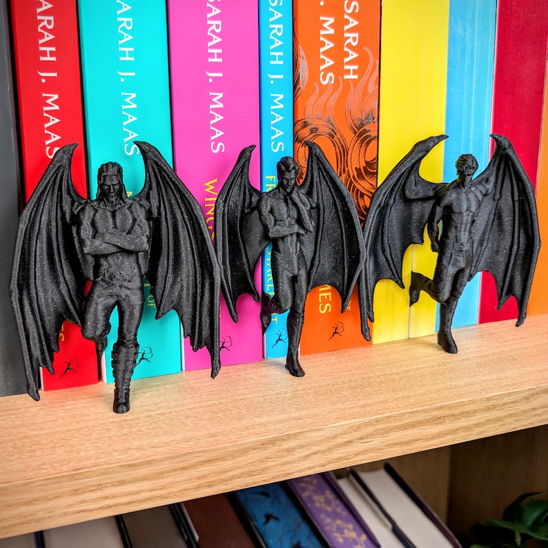 Puede incluir: Tres figuras negras impresas en 3D de personajes masculinos alados en diferentes poses. Cada figura presenta grandes y detalladas alas similares a las de un murci&eacute;lago y se encuentra de pie. Las figuras est&aacute;n sobre un estante de madera frente a libros.