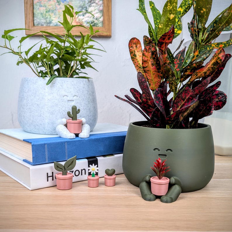 May include: Two decorative plant pots with smiling faces, one light gray and one olive green, each holding a small pink pot with a plant. Additional small pink pots with plants are in front of the pots. The pots are on a wooden surface.