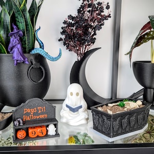May include: A black Days Til Halloween countdown calendar with orange blocks, a white ghost figurine reading a book, and a black coffin-shaped planter containing a skeleton. The set includes a purple witch figure and a black crescent moon.