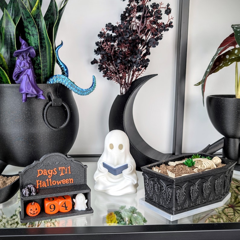 May include: A black Days Til Halloween countdown calendar with orange blocks, a white ghost figurine reading a book, and a black coffin-shaped planter containing a skeleton. The set includes a purple witch figure and a black crescent moon.