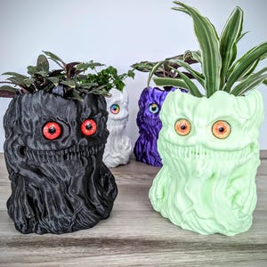 Puede incluir: Cuatro macetas de diferentes colores con forma de monstruo con ojos rojos y una sonrisa de dientes. Las macetas están hechas de plástico impreso en 3D y están llenas de plantas.