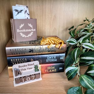 Peut inclure: Une pr&eacute;sentation de livres et d'accessoires. Une figurine de dragon dor&eacute; est pos&eacute;e sur trois livres, dont "Fourth Wing" et "Onyx Storm". Une petite bo&icirc;te marron avec l'inscription "Bookmarks" et un compteur "Books Read This Year" sont &eacute;galement pr&eacute;sents.