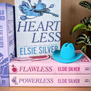 May include: A bright turquoise miniature cowboy hat resting on a stack of three books titled Heartless, Flawless, and Powerless by author Elsie Silver. The books feature light pink and white covers with floral illustrations and bold text.