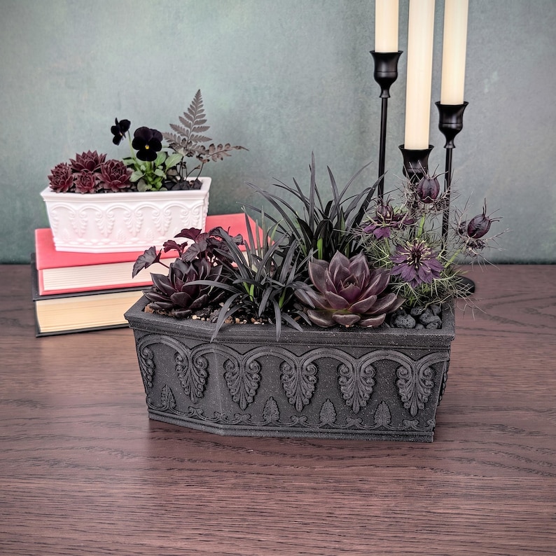 K&ouml;nnte beinhalten: Ein schwarzer rechteckiger Pflanzk&uuml;bel mit ornamentalen Reliefdetails, der dunkle Sukkulenten und violette Blumen enth&auml;lt, neben einem B&uuml;cherstapel mit einem wei&szlig;en Pflanzgef&auml;&szlig; mit dunklem Laub und Blumen sowie einem schwarzen dreiarmigen Kerzenst&auml;nder.