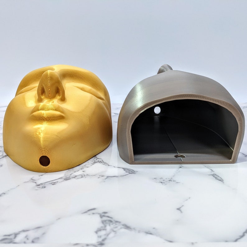 May include: A gold-coloured 3D printed face with closed eyes and a small hole in the bottom. Next to it is a grey 3D printed object with a hollowed-out interior.