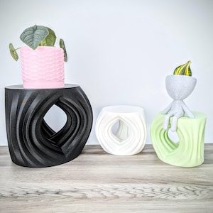 May include: Three black, white, and green 3D printed side tables with a unique wave design and a hole in the center. A pink planter with a plant is on the black table. A small white figurine with a green gourd on its head is sitting on the green table.
