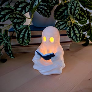 May include: A white, ghost-shaped lamp with a smooth, matte finish. The figure has two hollow, oval-shaped eye openings that emit a warm, glowing yellow light. The ghost is holding a small, dark-colored, open book in its hands.