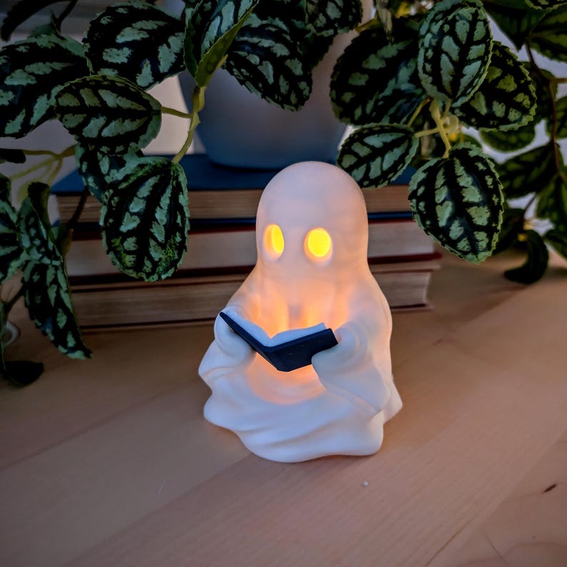 May include: A white, ghost-shaped lamp with a smooth, matte finish. The figure has two hollow, oval-shaped eye openings that emit a warm, glowing yellow light. The ghost is holding a small, dark-colored, open book in its hands.