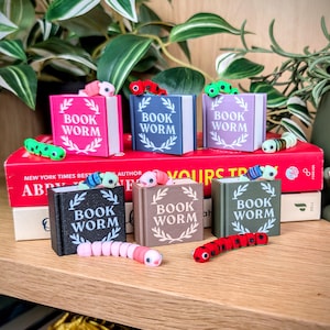 Puede incluir: Una colección de cajas en miniatura con forma de libro con el texto "BOOK WORM" en las portadas, cada una con una pequeña figura de oruga colorida. Los libros son de varios colores, incluyendo rosa, azul y negro, y están colocados en una estantería de madera.