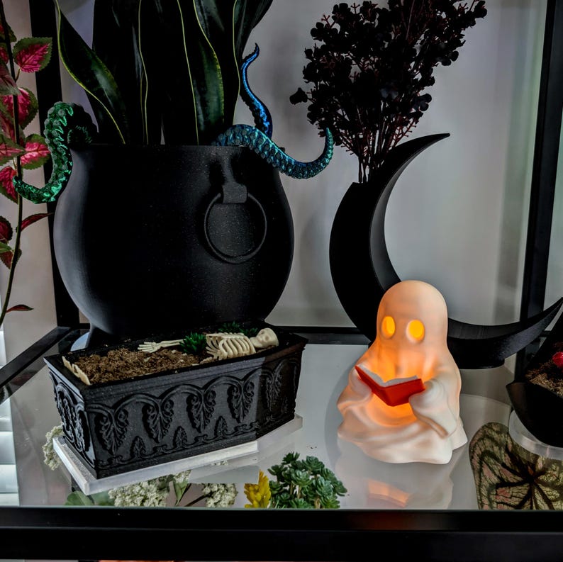 May include: A black cauldron planter with blue tentacles, a black coffin-shaped planter containing a skeleton, and a small white ghost figurine holding a red book. The ghost is illuminated from within, casting a warm glow on the red book.