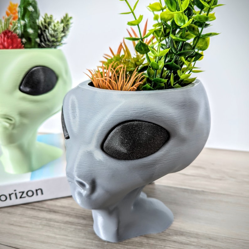 May include: A gray alien-shaped planter with a green plant inside. The planter has a large, black eye and a small, black nose.