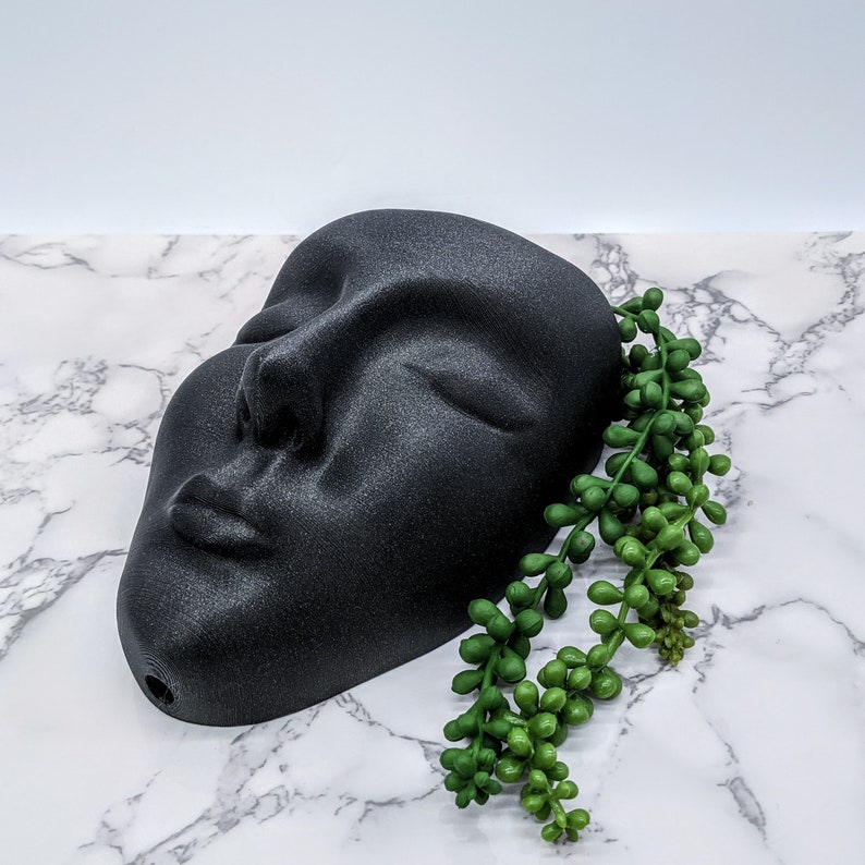 May include: A black face planter with a closed eye and mouth, resting on a white marble surface. A green succulent plant is placed next to the planter.