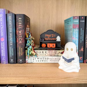 May include: A black Days Til Halloween countdown sign with orange blocks displaying the number 18, a small white ghost figure, and an orange pumpkin. A metallic iridescent witch figurine stands nearby, and a white ghost figurine holds a blue book.