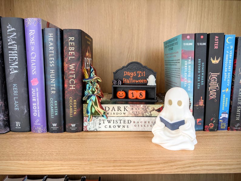 May include: A black Days Til Halloween countdown sign with orange blocks displaying the number 18, a small white ghost figure, and an orange pumpkin. A metallic iridescent witch figurine stands nearby, and a white ghost figurine holds a blue book.