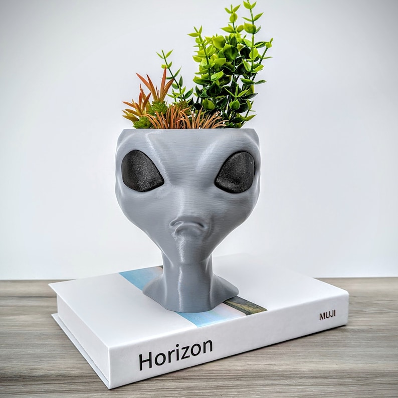 May include: A gray alien-shaped planter with black eyes, holding a small potted plant with green leaves. The planter is sitting on a white book with the word "Horizon" printed on the cover.