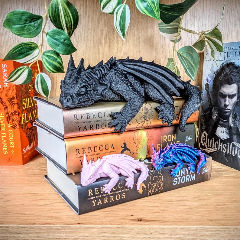 Puede incluir: Una figura de drag&oacute;n negro descansa sobre una pila de libros con el nombre de la autora, Rebecca Yarros, visible. Tambi&eacute;n hay dos figuras de drag&oacute;n m&aacute;s peque&ntilde;as, una rosa y otra azul.