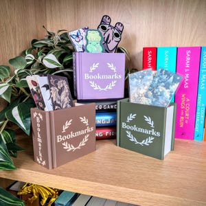 May include: Three book-shaped containers in brown, olive green, and lavender, each labeled "Bookmarks" with a laurel wreath design. The containers hold various decorative bookmarks. The background includes books and greenery.