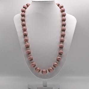 Puede incluir: Un collar de cuentas rosas expuesto sobre un maniquí blanco. El collar presenta grandes cuentas texturizadas ensartadas. Las cuentas tienen un aspecto moteado y son de un color rosa empolvado. El collar es una pieza llamativa.