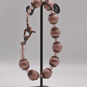 Puede incluir: Una pulsera de cuentas rosas con cuentas redondas texturizadas y cuentas facetadas más pequeñas. La pulsera presenta un cierre de color cobre y se exhibe en un soporte negro. Las cuentas tienen un diseño detallado en relieve.