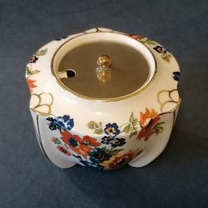 May include: A white ceramic sugar bowl with a floral design in shades of blue, orange, and yellow. The bowl has a gold trim and a lid with a gold knob.