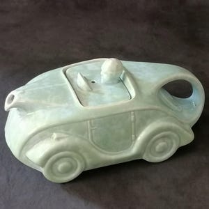 May include: A pale green ceramic teapot in the shape of a classic car. The teapot has a driver figure, a spout, a handle, and four wheels. This car-shaped teapot is a unique and decorative kitchen item.