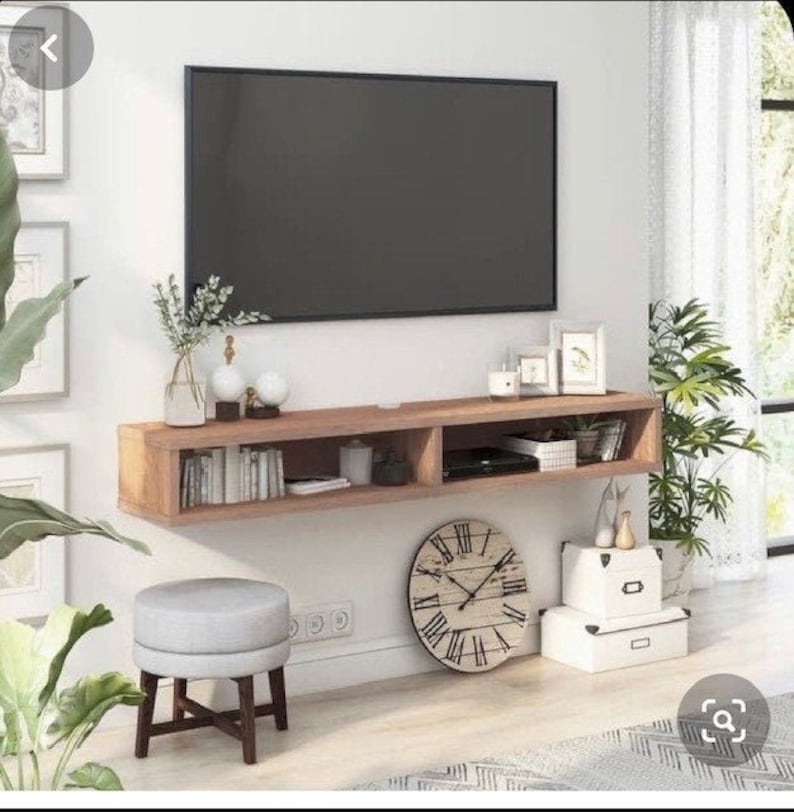 May include: A modern living room setup featuring a large black flat-screen TV mounted above a wooden floating shelf. The shelf holds books, decor, and a media player. A gray stool, a large clock, and plants complete the room's design.