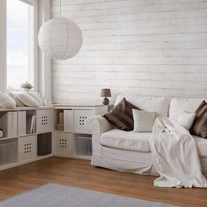 May include: A white couch with brown throw pillows and a white blanket sits in a living room with whitewashed wood paneling on the wall. The room has a white ceiling and a large window with a view of the sky. There is a white lamp on a side table next to the couch.