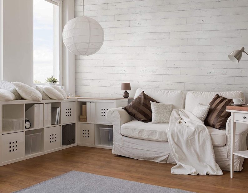 May include: A white couch with brown throw pillows and a white blanket sits in a living room with whitewashed wood paneling on the wall. The room has a white ceiling and a large window with a view of the sky. There is a white lamp on a side table next to the couch.